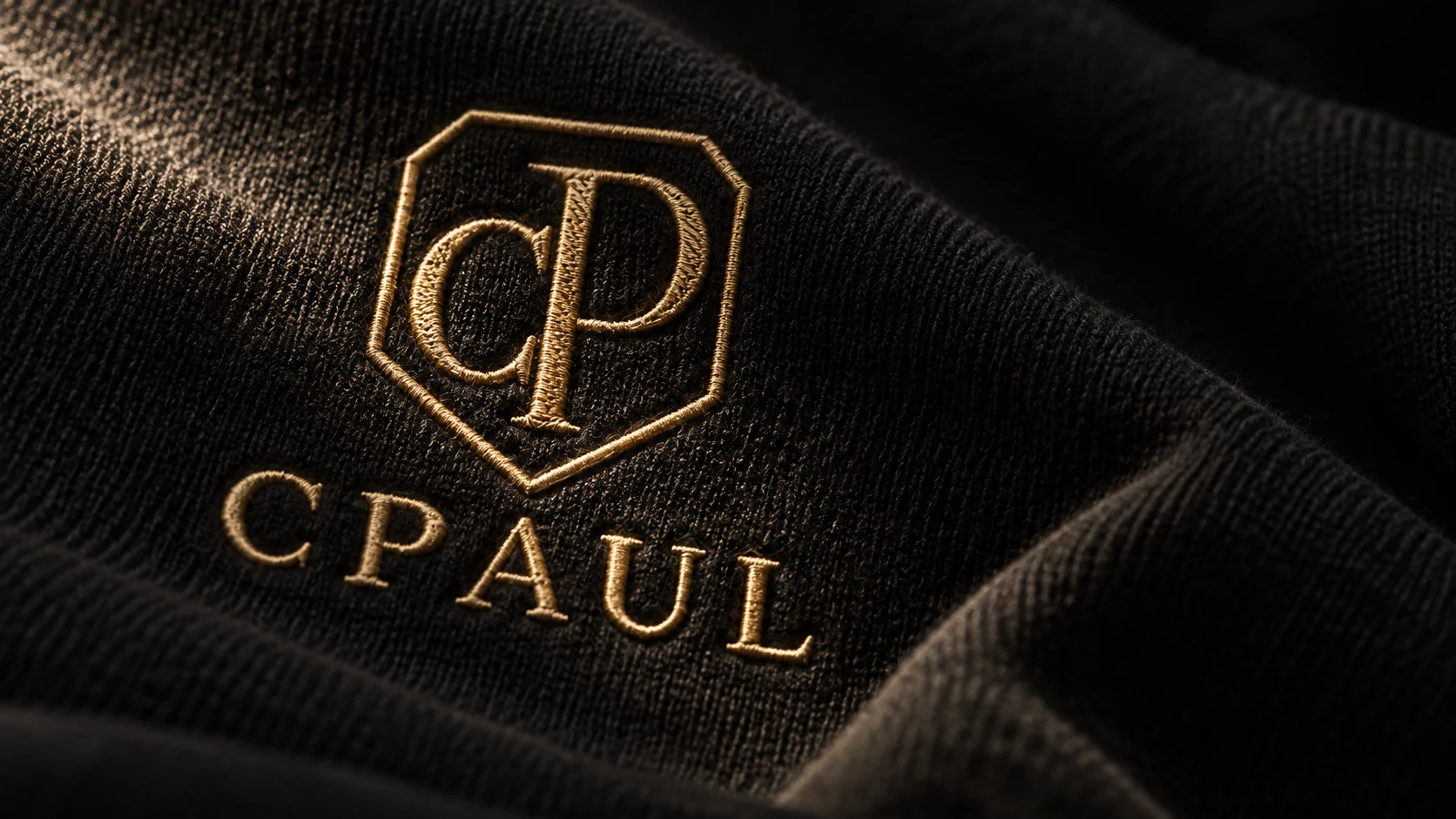 Imagen de campaña: detalle de bordado dorado CPAUL sobre una tela negra